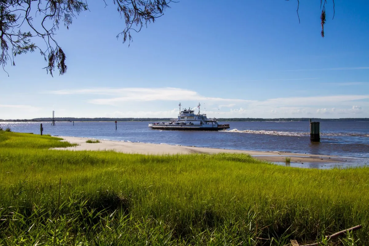 A serene view of the Southport-Fort Fisher ferry crossing a tranquil waterway, bordered by lush green grass and with a distant shoreline visible. The ferry is the focal point, inviting thoughts of day trips and local exploration.