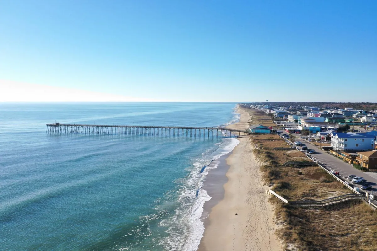 A broad view of Kure Beach Pier stretching over the ocean, with the sandy beach and shoreline in the foreground and houses lining the coast. The area is bathed in natural light, creating a lively and inviting atmosphere ideal for group adventures.