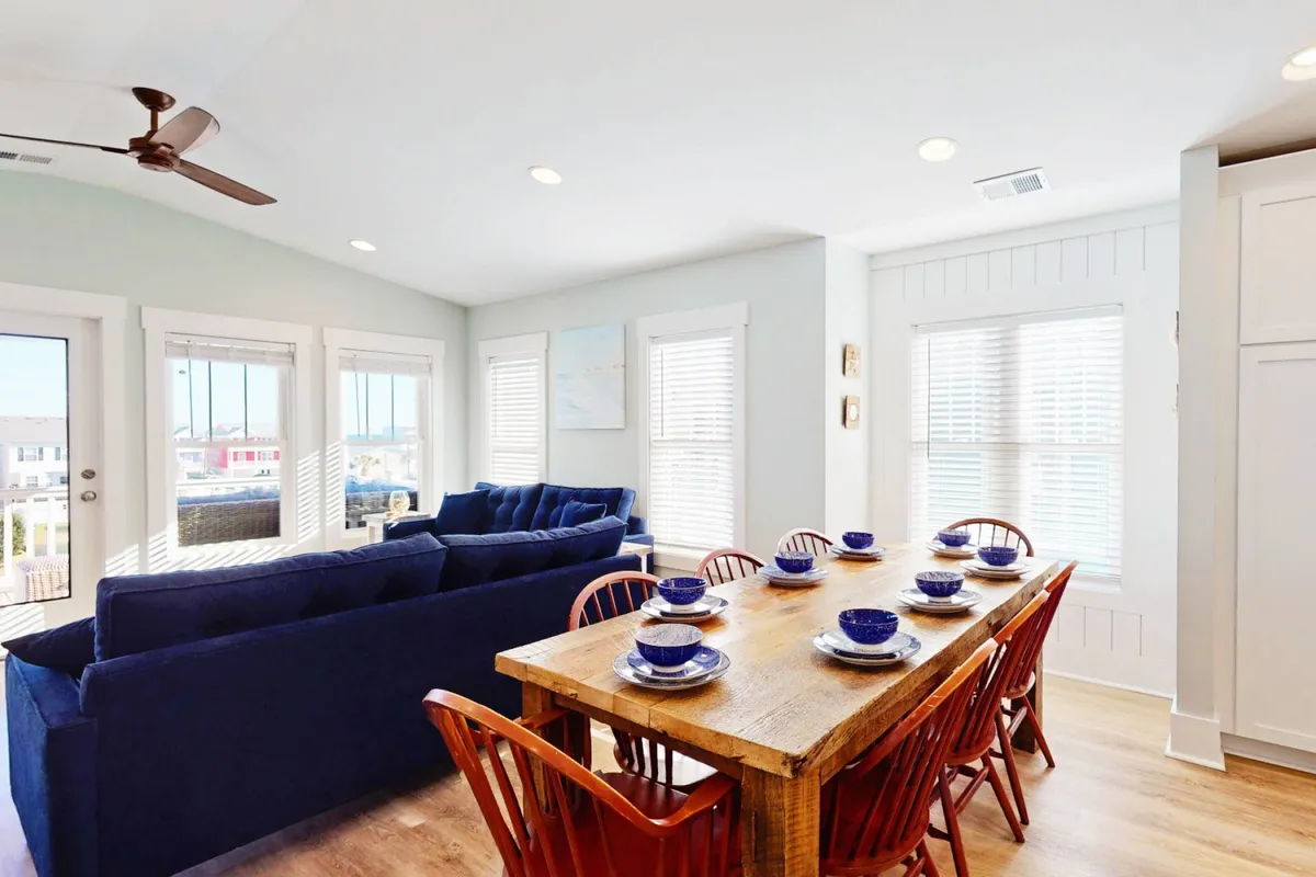 A bright dining space highlighted by a substantial dining table, blue sofa seating, and a window that frames the outside view. The space is designed for group meals, celebrations, and casual gatherings.