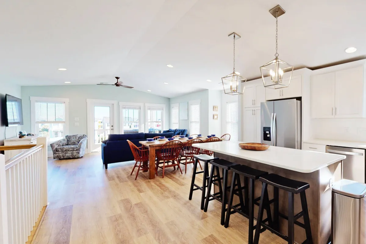 This image displays an open-concept living area that includes a spacious island countertop, a dining table suitable for gatherings, comfortable sofa seating, and large windows that flood the space with natural light. The windows appear to offer a glimpse of the outdoors, reinforcing the sense of openness and connection to the surrounding environment.