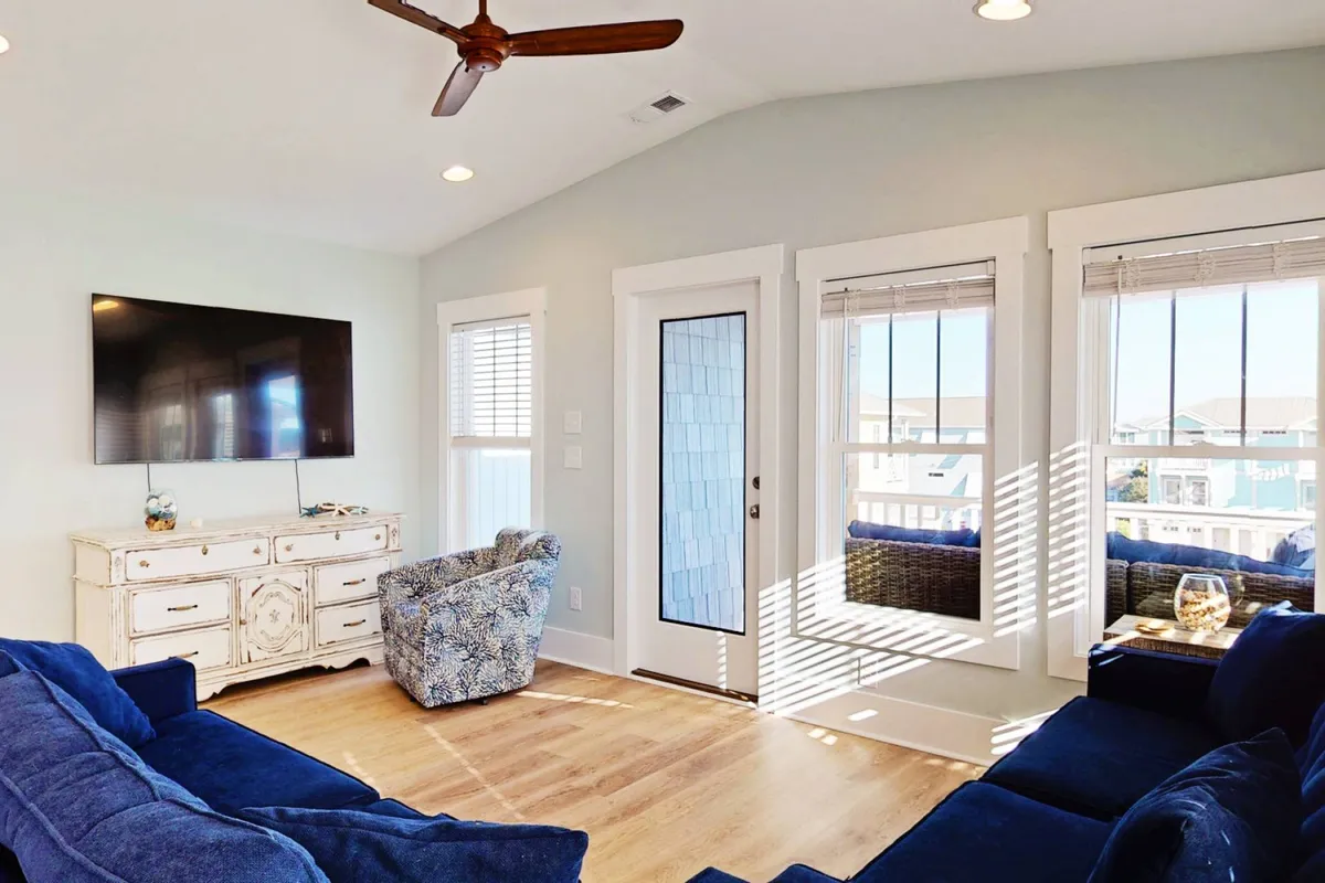 A cozy living area with a blue sectional sofa, decorative armchair, wall-mounted TV, decorative console table, and large windows streaming in natural light. The setup invites relaxation, socializing, and entertainment, with views outward that connect the interior to the exterior setting.