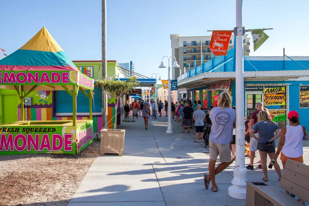 A bustling boardwalk scene with a colorful lemonade stand, several bright storefronts, crowds of people, lampposts, and benches. The mood is lively and festive, suggesting opportunities for socializing and sampling local treats.