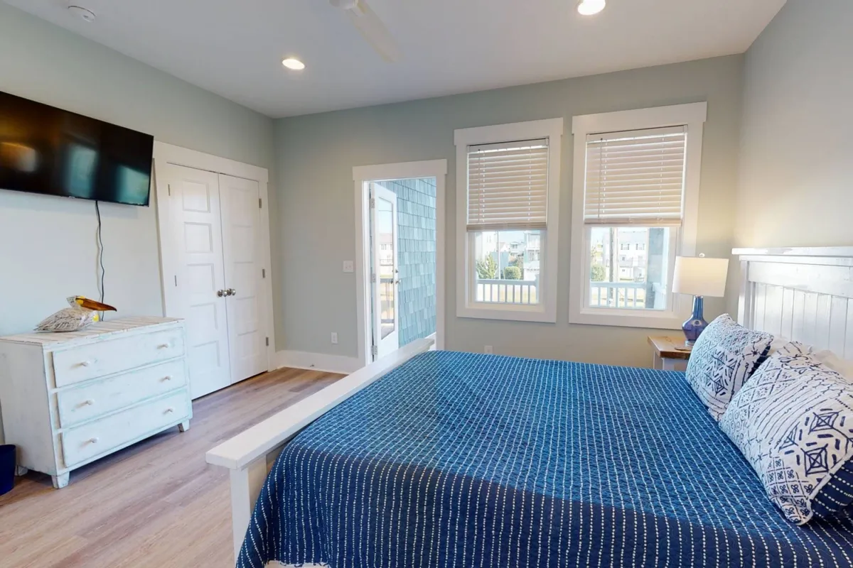 A tranquil bedroom featuring blue patterned bedding, a television mounted on the wall, large windows with blinds letting in natural light, and a wooden dresser. The space is inviting and designed for comfort and relaxation.