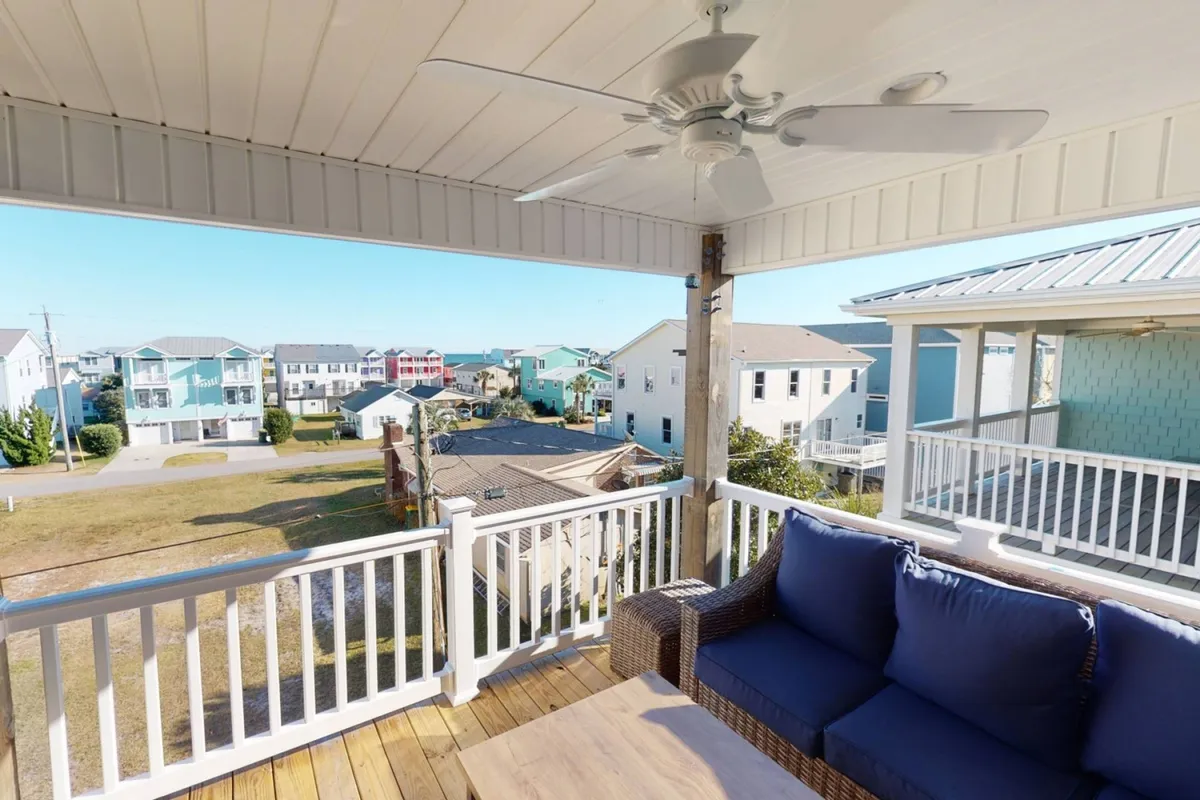 The image showcases a serene outdoor seating area on a balcony, perfect for relaxing and socializing, with sweeping views of neighboring coastal houses, a calm skyline, and the sparkling ocean visible in the distance. The scene evokes a tranquil, inviting atmosphere ideal for sunrise coffee or evening conversations while gazing out over Kure Beach.