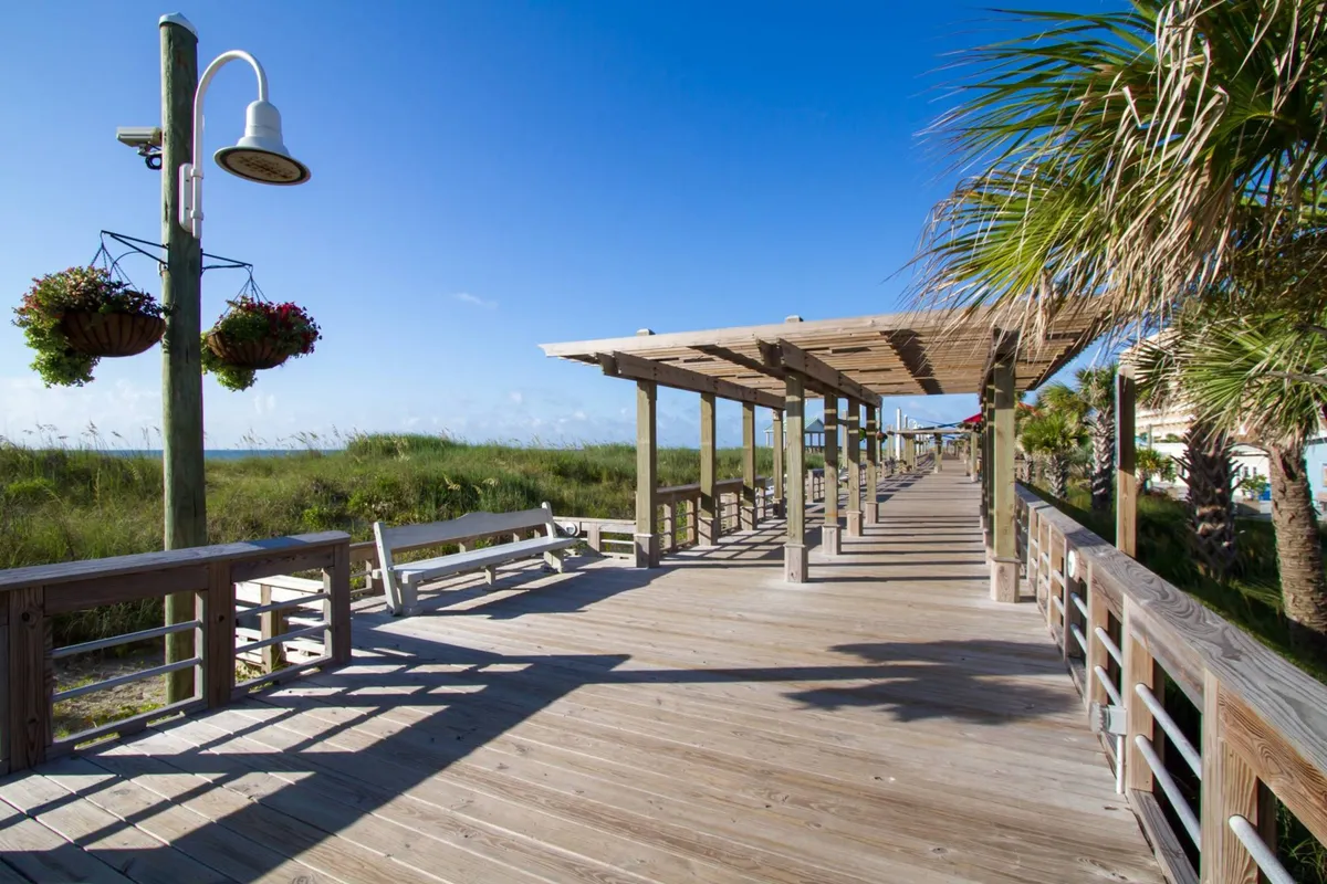 A peaceful exterior scene featuring a wooden boardwalk lined with hanging flower baskets and palm trees, set under an open sky. The scene is inviting and suggests leisurely strolls and outdoor relaxation.