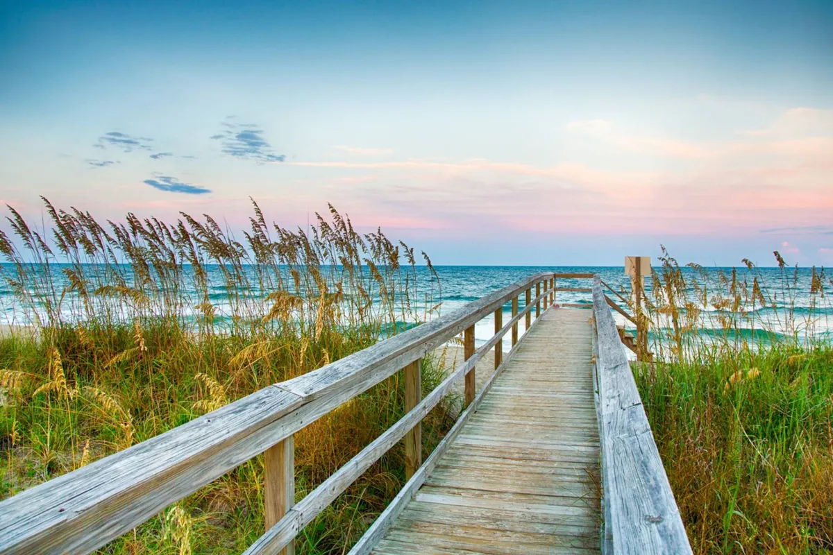 A tranquil view from a wooden boardwalk overlooking the ocean, with dune grass in the foreground and pastel-colored skies above. The mood is calm, perfect for unwinding after a day of adventures.