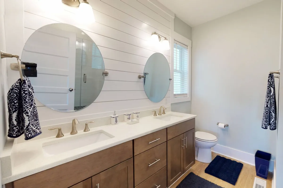A modern bathroom featuring dual round mirrors, modern light fixtures, a wood-finished vanity, and a pristine white countertop. The setting is both inviting and tranquil, perfect for morning routines or evening unwinding.