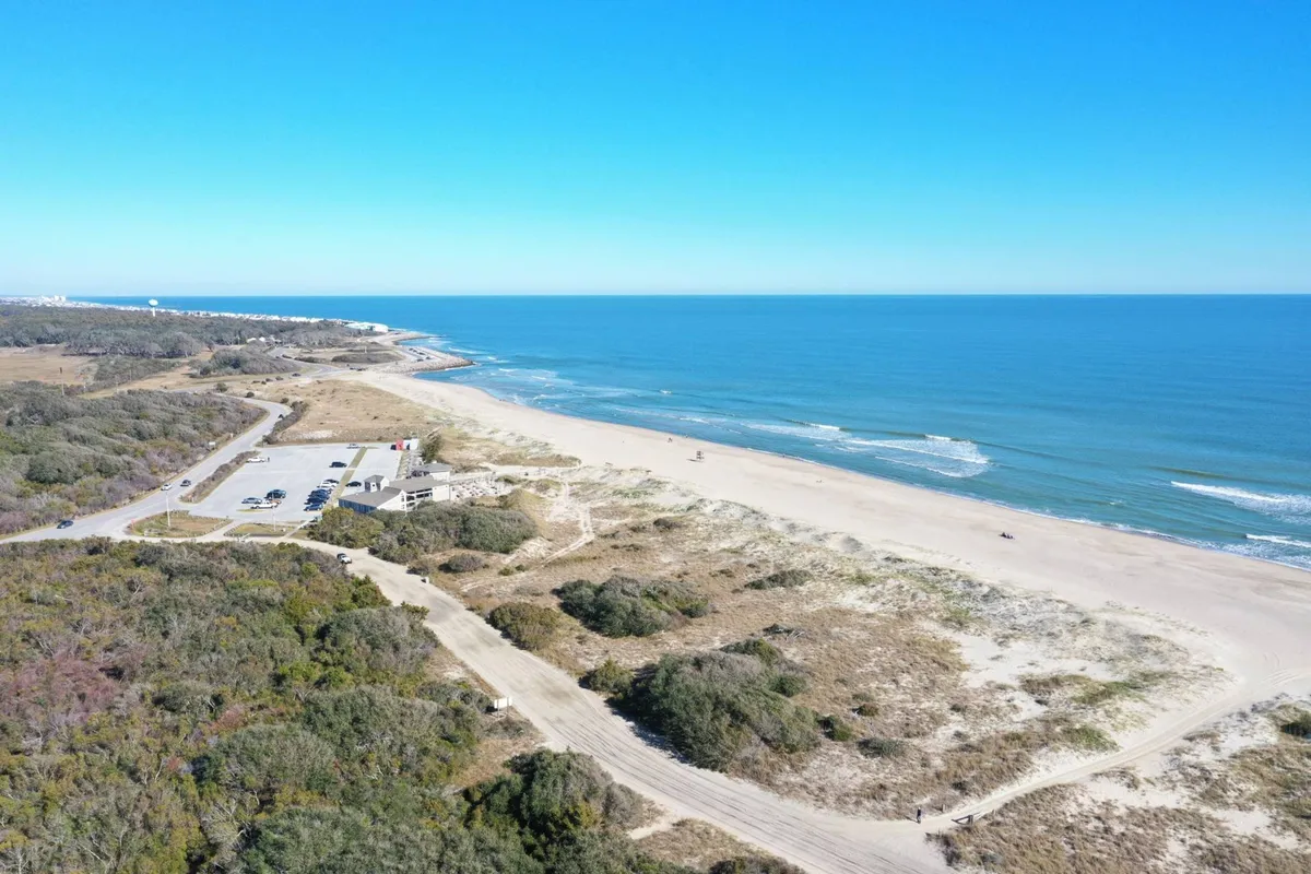 A panoramic exterior scene of the beach and ocean, with parking areas, sandy dunes, and lush coastal vegetation visible. The view suggests easy access for groups and highlights the stunning natural setting.
