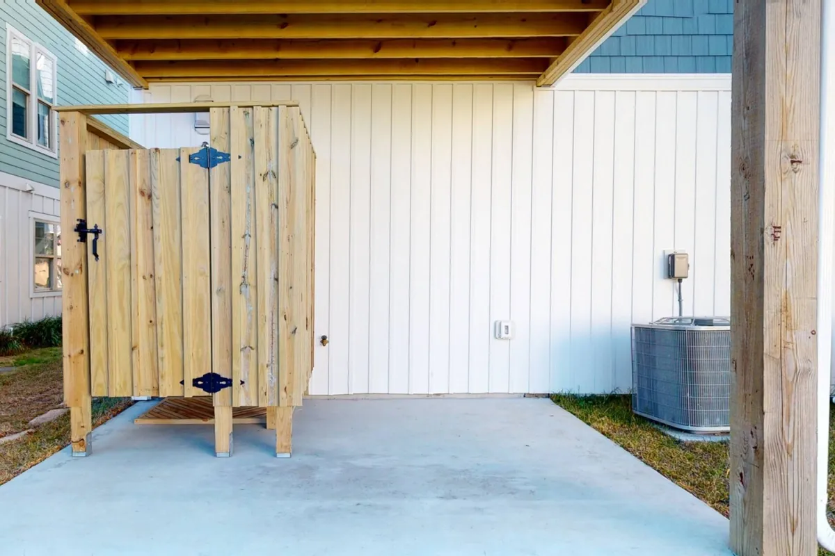 An exterior shot of a wooden outdoor shower enclosure sits beside a clean, shaded concrete area, with an air conditioning unit visible nearby. The setup is practical, inviting, and ideal for quick rinses after beach time.